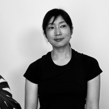 Portrait of Sachi Hasegawa. A woman with her hair tied back. Black-and-white photograph with a leaf of a houseplant on the left.