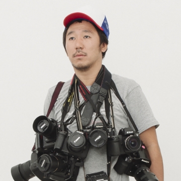 Portrait of Masanori Ikeda. A man with a cap and a beard. He has many cameras and lenses hanging from his neck.