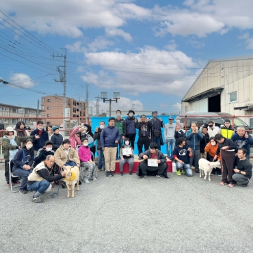 Members of Mugiwaraya. Outdoors on a sunny day, about 30 people are lined up in a semicircle with goats and other animals.